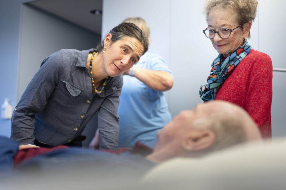 Palliaviva-Mitarbeiterin Livia de Toffol (links) beugt sich zu einem Patienten, der im Bett liegt. Daneben steht seine Frau. Foto: Gaëtan Bally